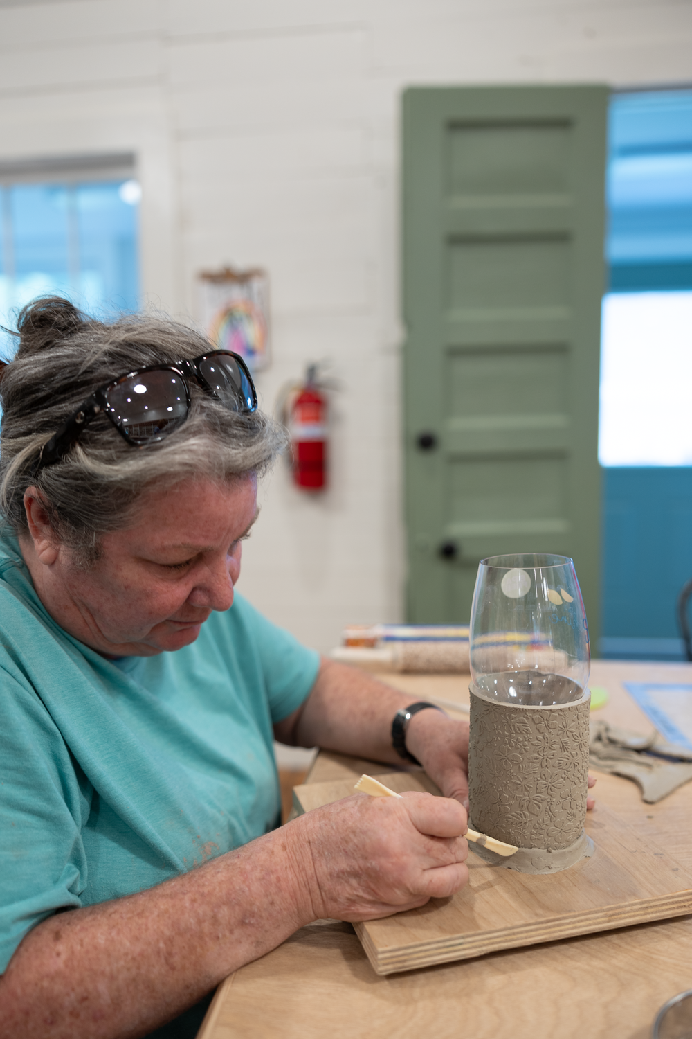 A craftsperson wearing a teal shirt carefully decorates a glass vase with sand art at a wooden workbench indoors.