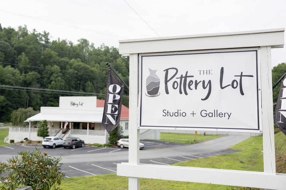 The Pottery Lot Studio and Gallery sign with open flag, white building and lush green trees in background.