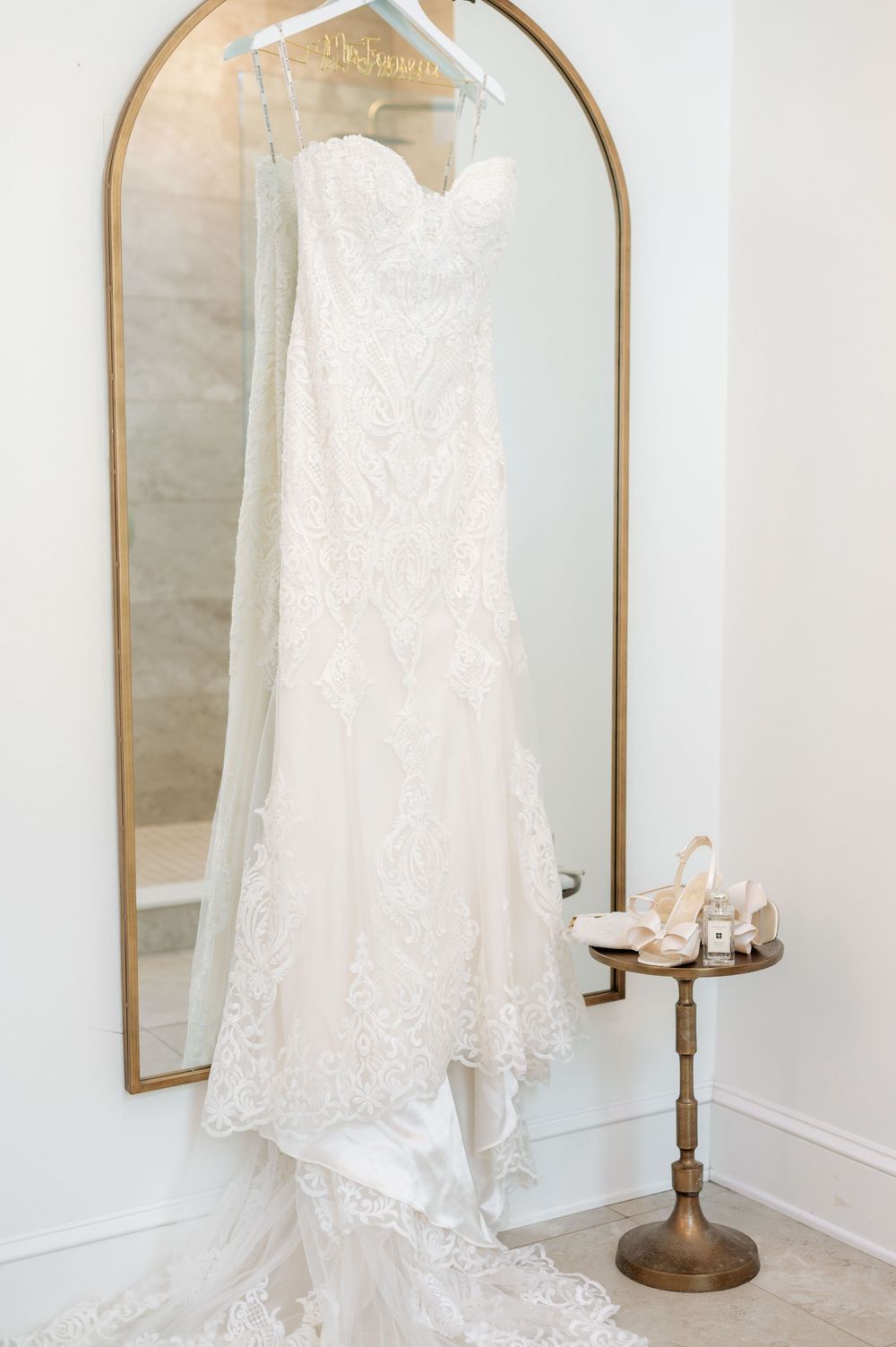 Elegant white lace wedding dress hanging on display against arched mirrors in a bridal suite.