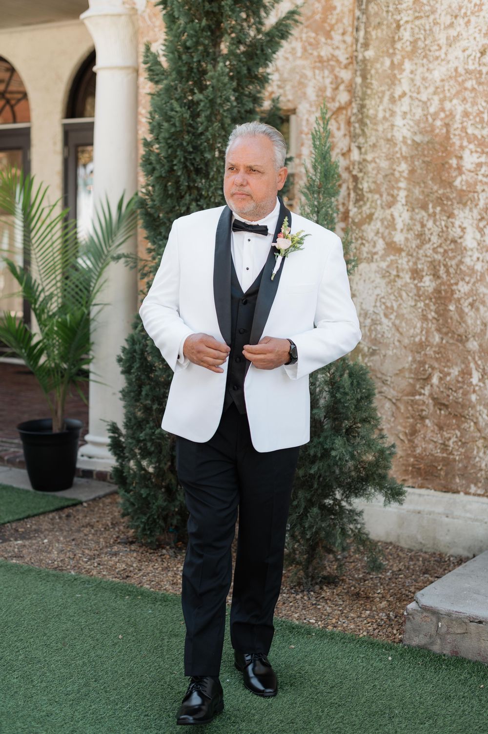Formal wedding attire featuring white dinner jacket, black bow tie and boutonniere in elegant venue setting.