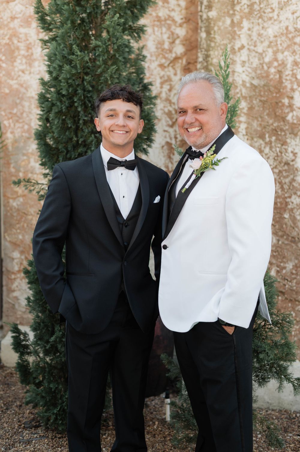 Formal wedding party members in black and white tuxedos pose against a textured wall with greenery.