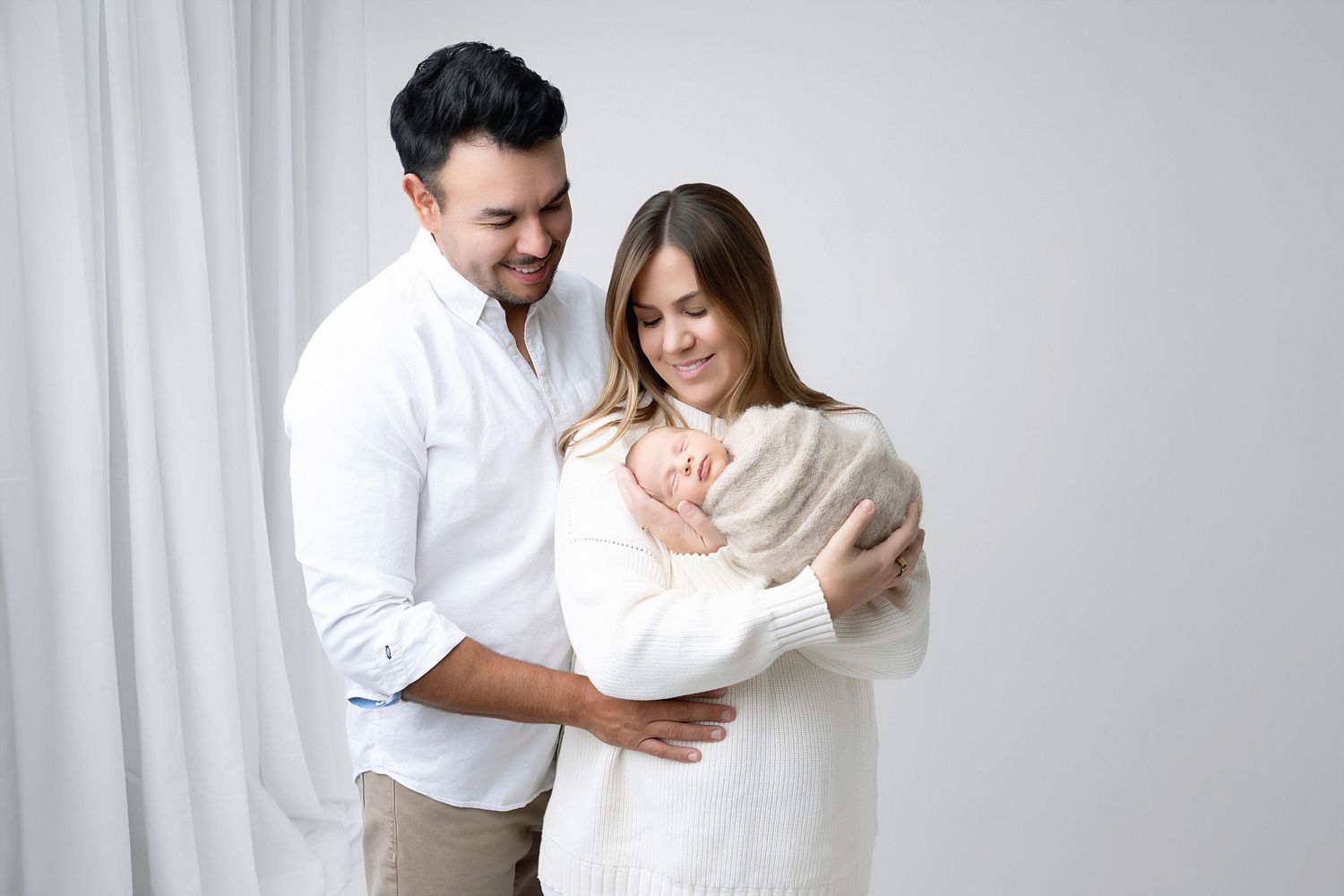 Expecting parents wrapped in white fabric embrace tenderly against light studio background.