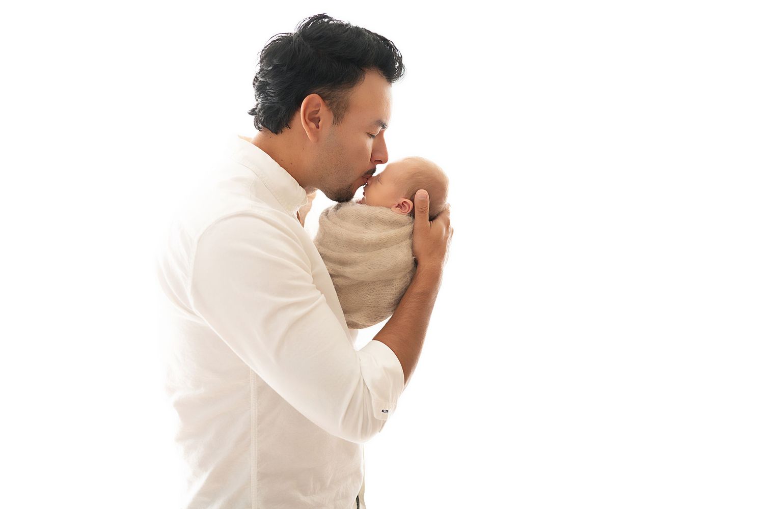 A person in a white shirt gently kisses a sleeping newborn baby wrapped in beige swaddling cloth.
