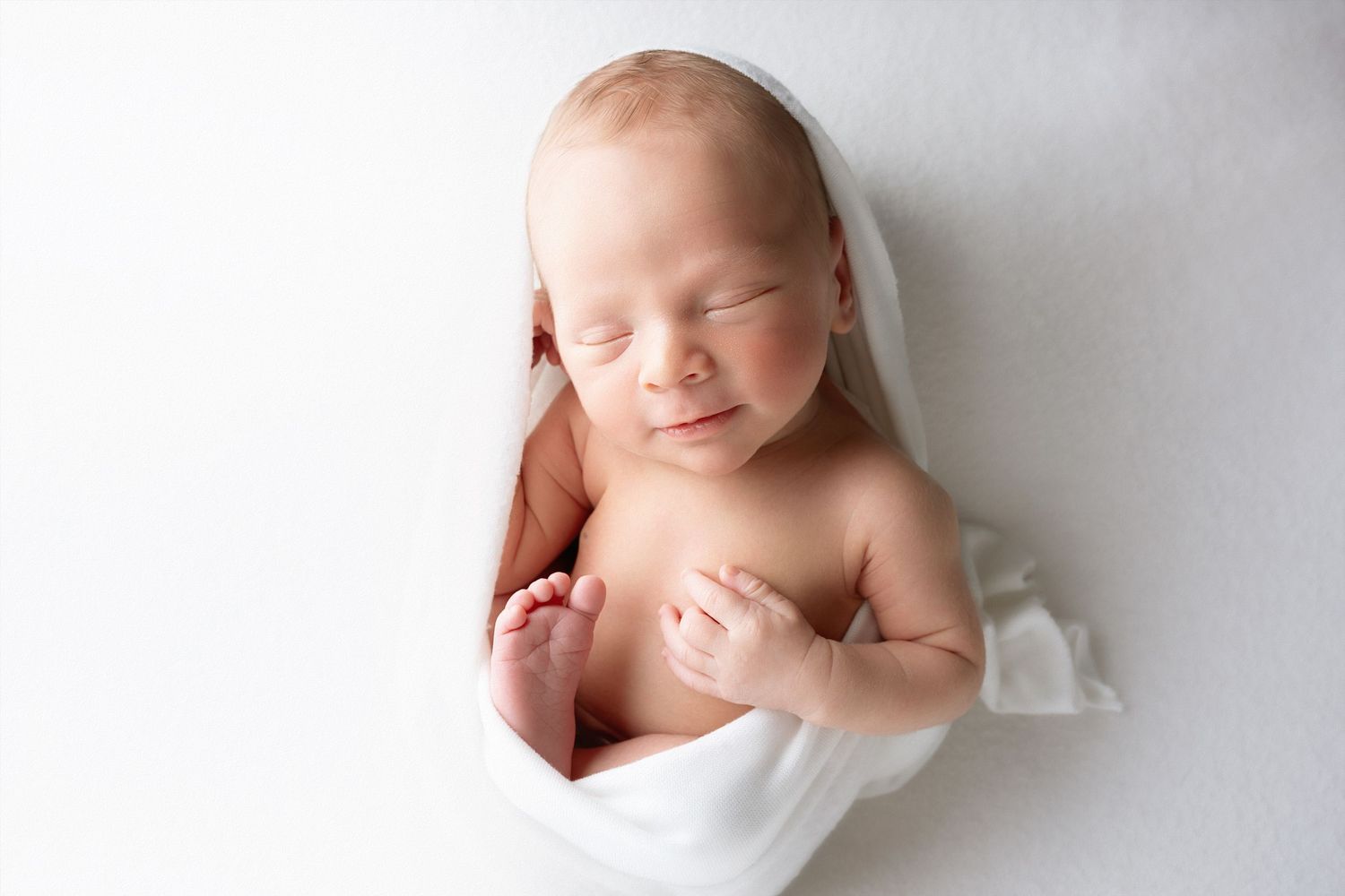 Peaceful sleeping newborn baby wrapped in soft white fabric against light background.