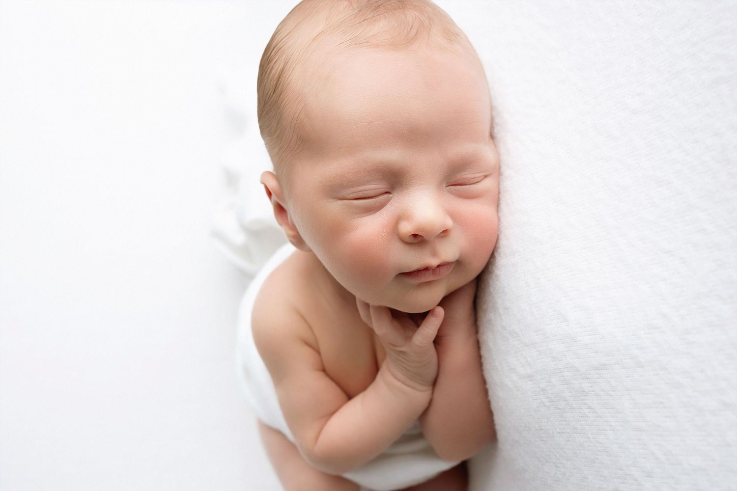 Peaceful sleeping newborn baby curled up on soft white backdrop in minimalist studio setting.