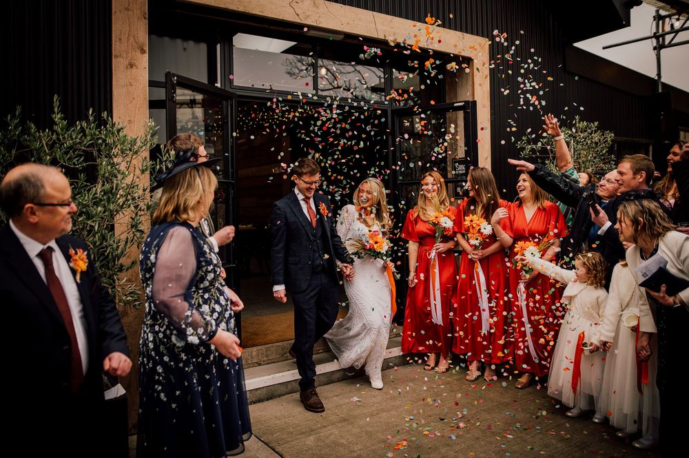 Wedding party members in red floral dresses and dark suits celebrate with confetti at an outdoor venue.