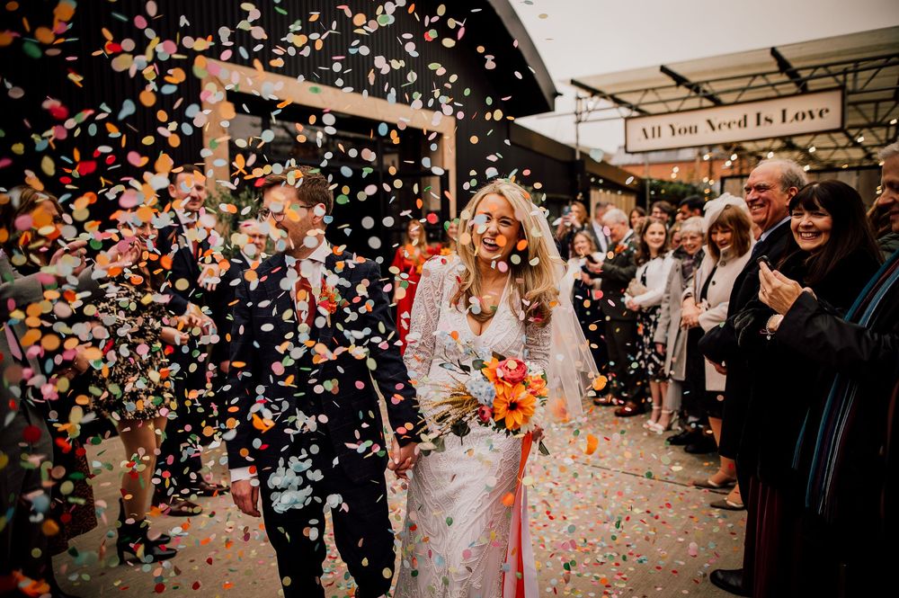 Newlyweds exit their wedding ceremony under a shower of colorful confetti while guests cheer and celebrate.