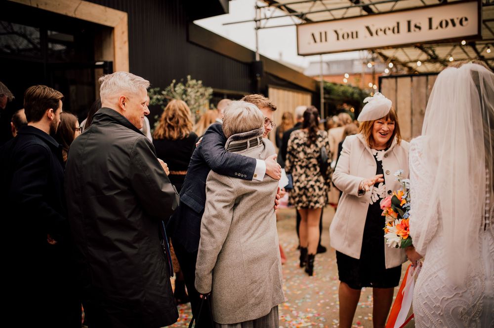 Wedding guests gather under a rustic 'All You Need Is Love' sign at an outdoor venue during a celebration.