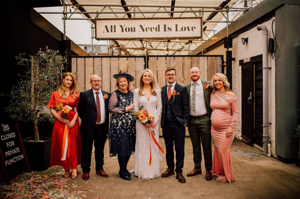 Wedding party poses beneath wooden 'All You Need Is Love' sign at rustic outdoor venue.