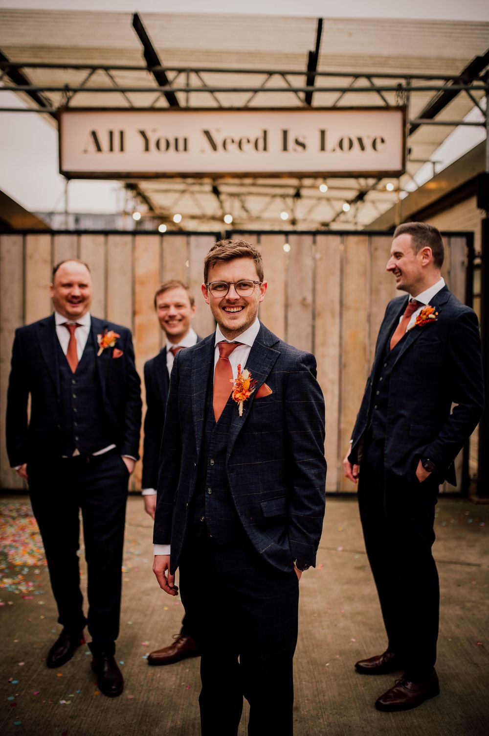 A groomsmen party in dark suits and orange ties pose playfully in front of a rustic industrial wedding venue wall.