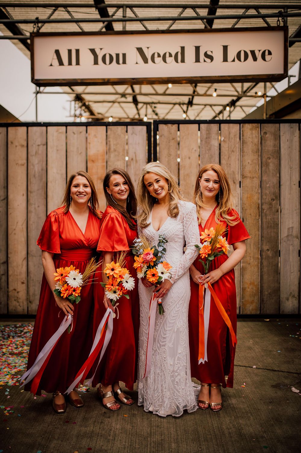 A bridal party poses under a rustic wooden sign reading 'All You Need Is Love' with bridesmaids in red dresses holding flowers.