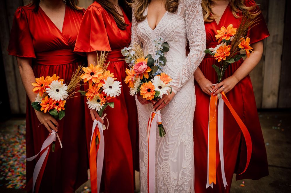Wedding party holds vibrant orange and white floral bouquets while wearing red and white dresses for autumn celebration.