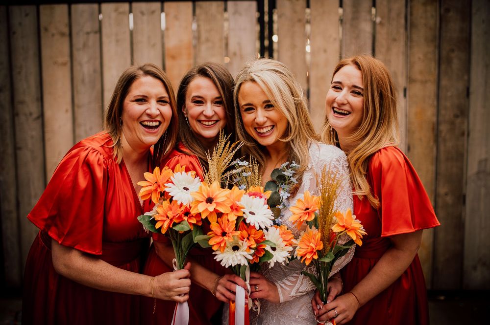 Bridesmaids in vibrant red dresses hold colorful orange and pink floral bouquets while smiling against wooden backdrop.