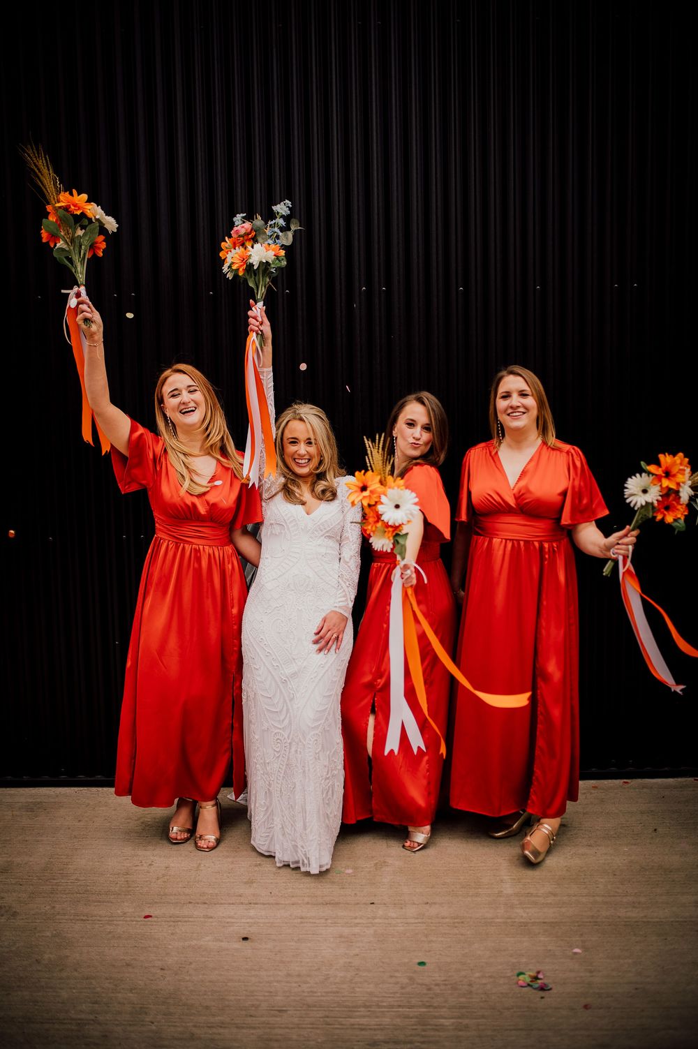 Wedding party in bright red dresses and white gown pose together against dark backdrop with orange floral bouquets.