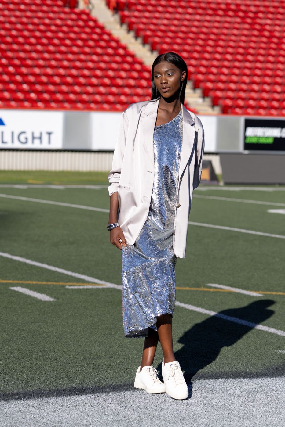 A person in a sparkly silver dress and white blazer poses on a football field with stadium seats in the background.