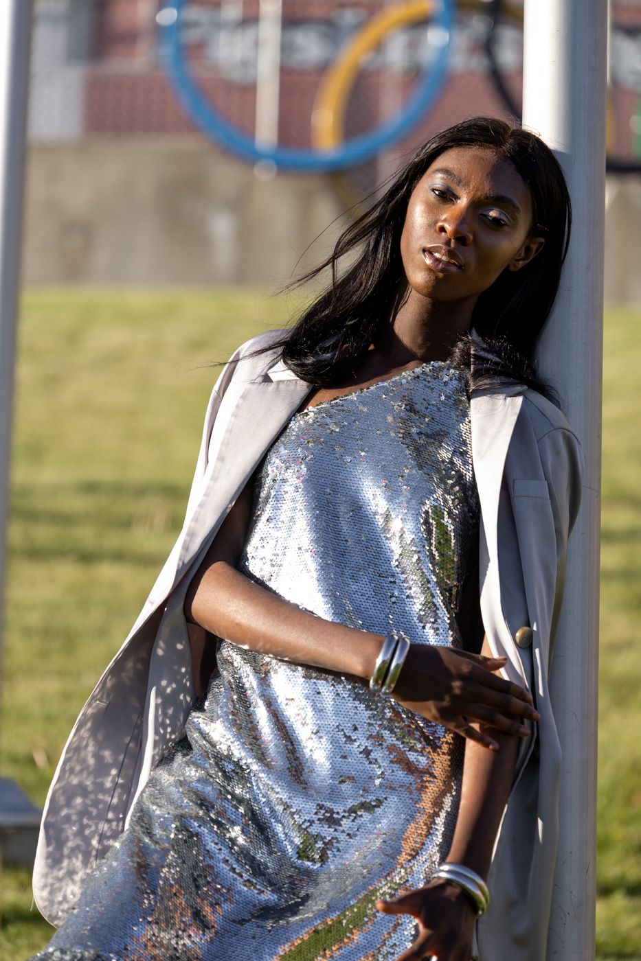 Model in shimmering silver dress and white blazer poses outdoors against playground equipment background.