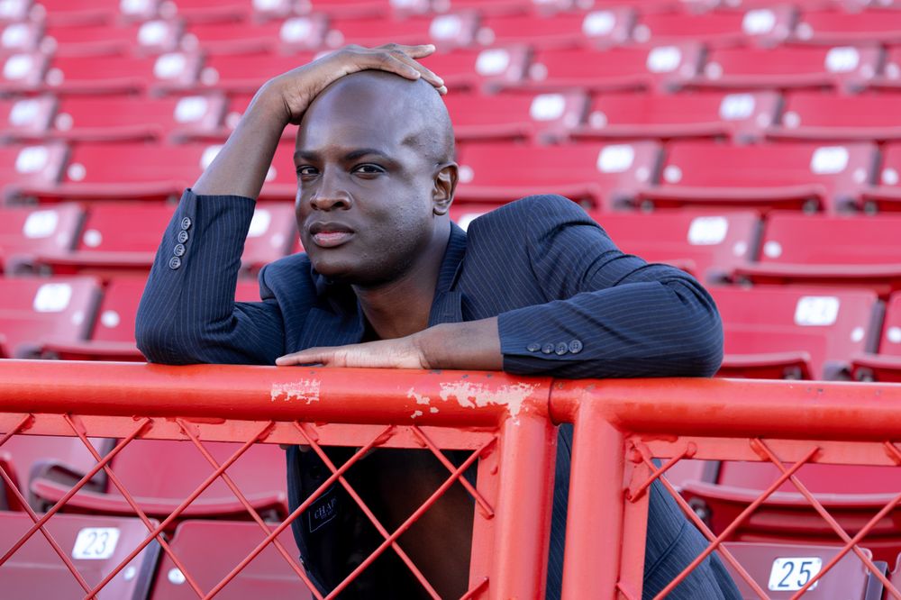 Professional in a dark suit jacket leans thoughtfully against red stadium seats at an empty sports venue.