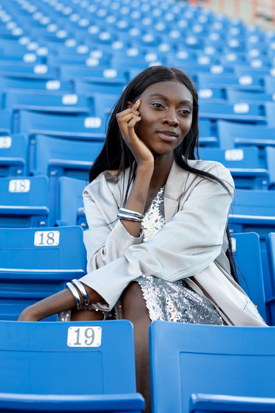 Person in white top poses thoughtfully in blue stadium seats during a photoshoot.