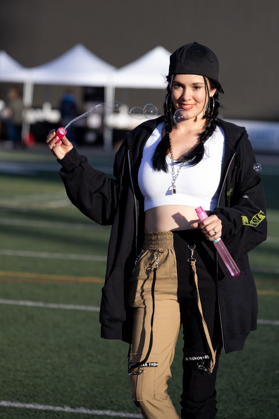 A person in a black jacket and khaki pants stands on an artificial turf field during a sunny day.