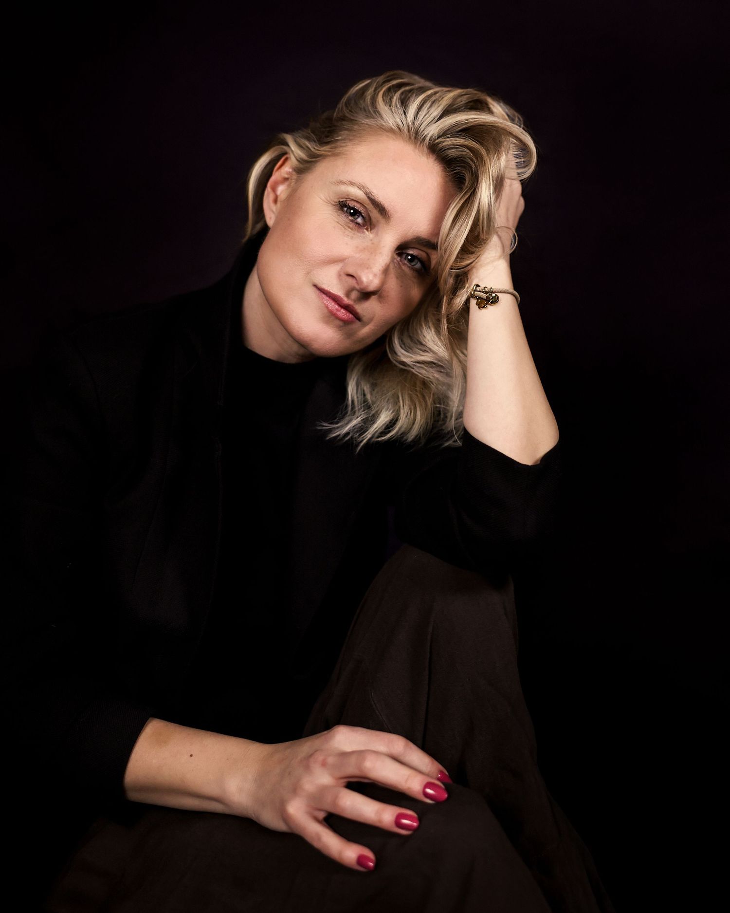 Portrait of a person in a black turtleneck sweater posing against dark background with blonde hair and red nail polish.