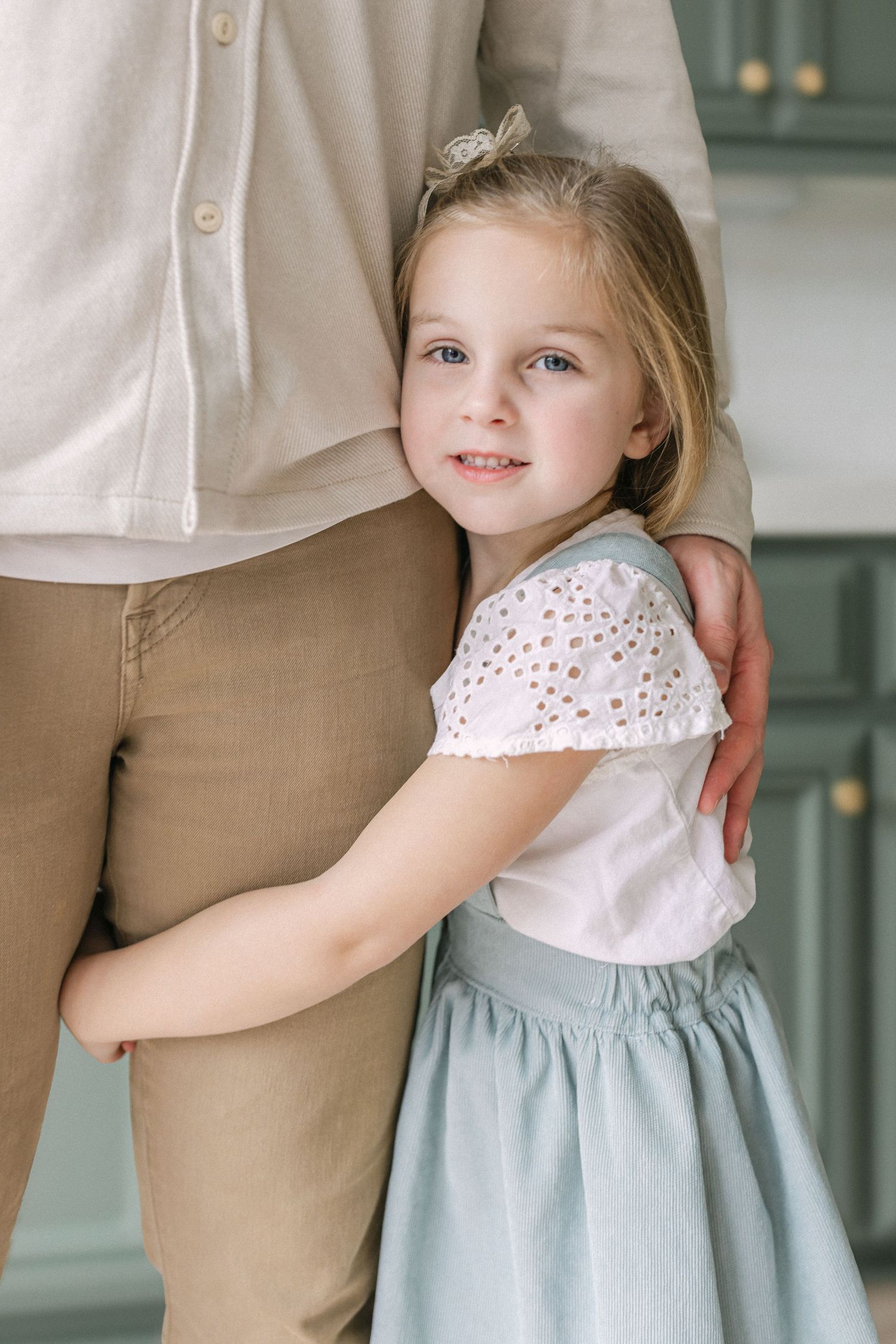 A young child in a blue and white dress hugs someone wearing khaki pants in a tender moment.