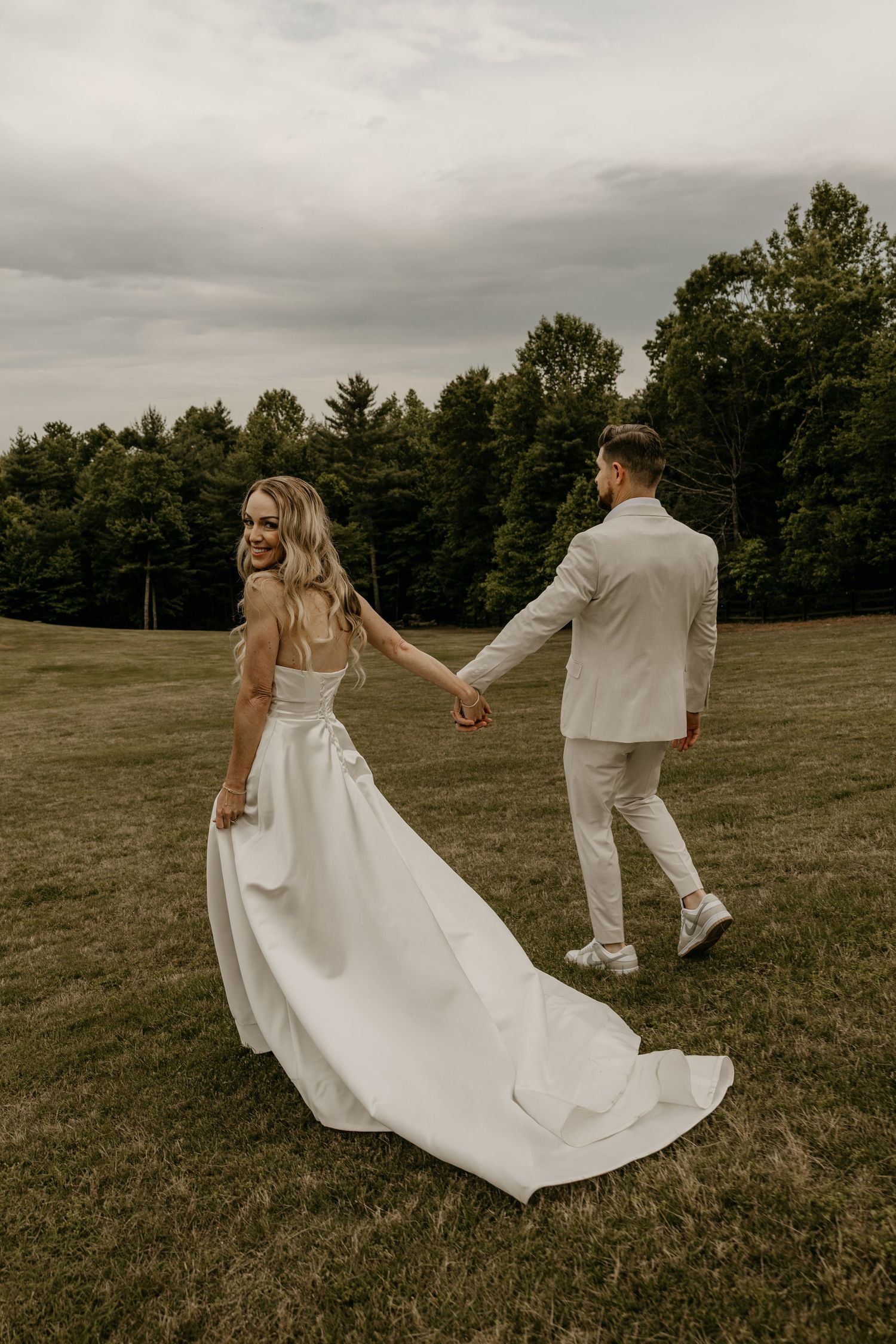 A couple in wedding attire holds hands and walks through a grassy field on an overcast day, the bride's dress flowing behind them.