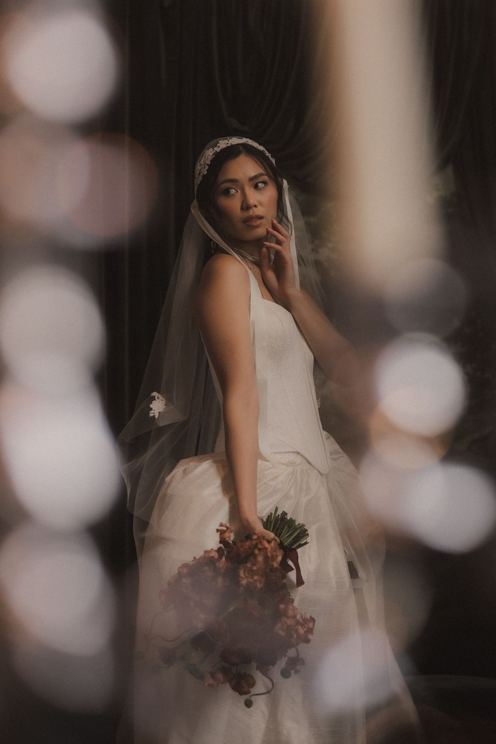 A bride in a classic white wedding dress and veil poses thoughtfully while holding a bouquet in moody lighting.