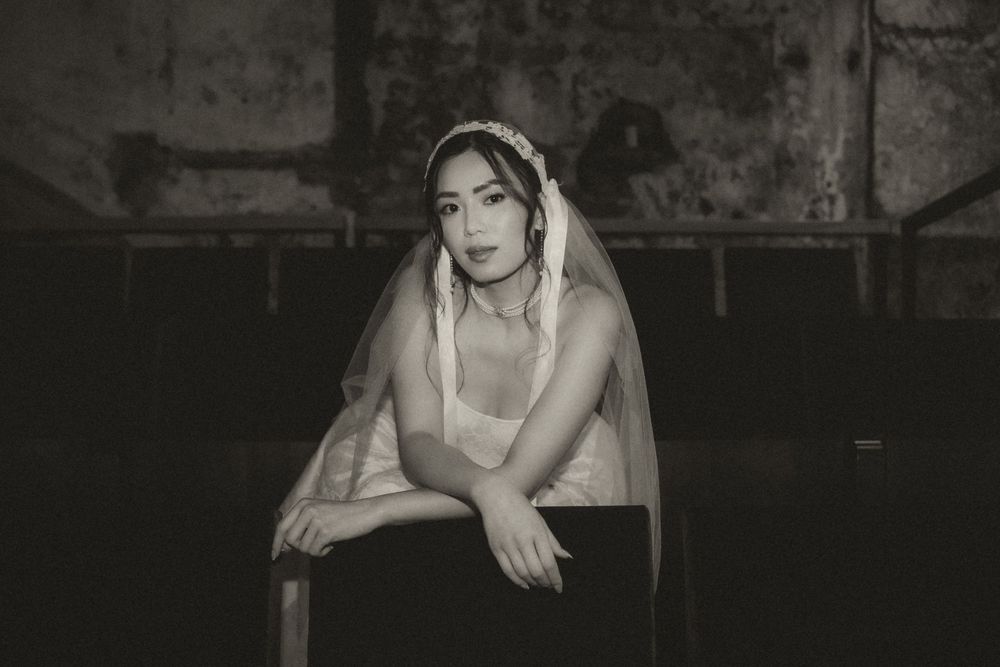 A black and white portrait of a bride in a wedding dress sitting thoughtfully in a dark, moody setting.