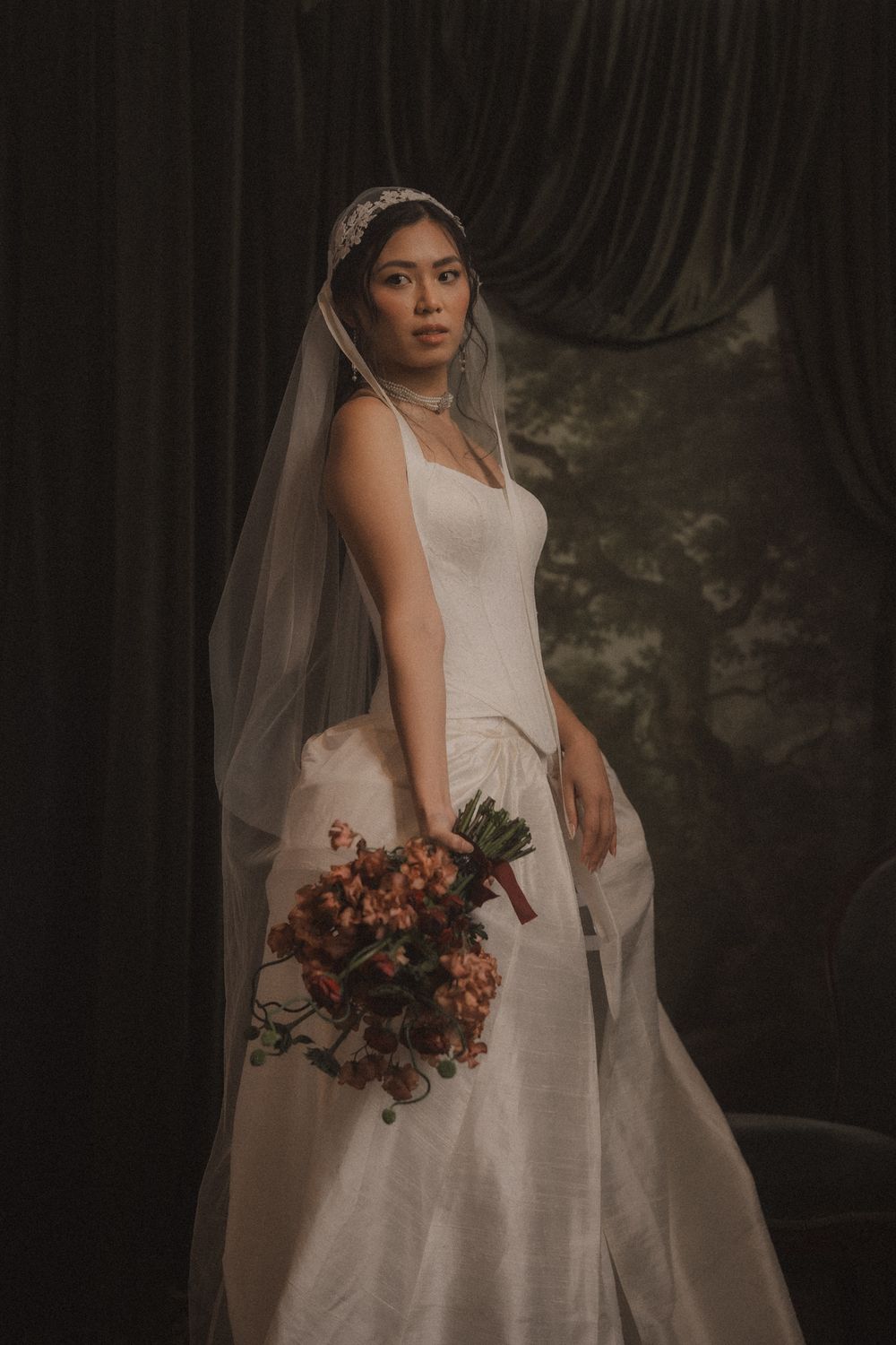 A bride in a classic white wedding dress holds an autumn-toned bouquet while posing against a dark dramatic backdrop.