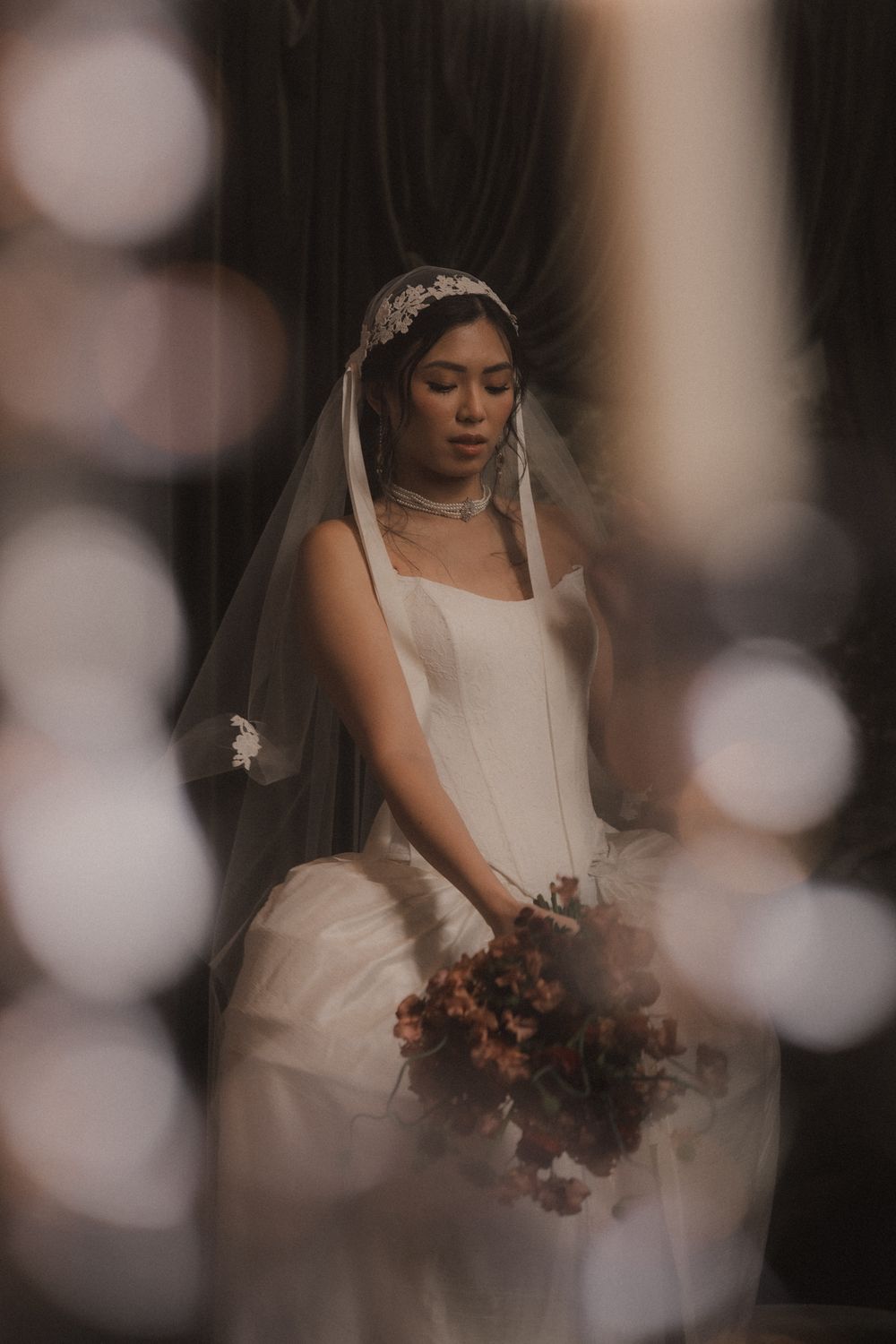 A bride in a classic white wedding dress and veil holds an autumn-colored bouquet in a dreamy, bokeh-filled setting.