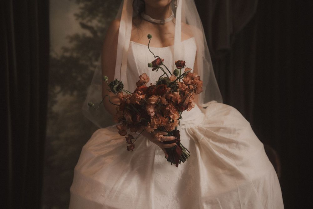 Bride in flowing white wedding dress holds autumn-colored bouquet against moody forest backdrop.