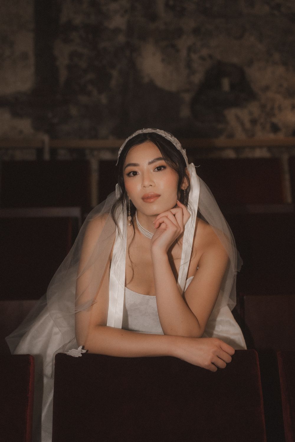 A bride in a white veil poses elegantly on a rustic wooden surface in a dimly lit setting.