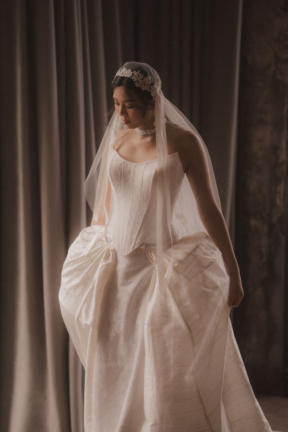 A flowing white wedding dress with long sleeves and veil is photographed against a dark background in an artistic style.