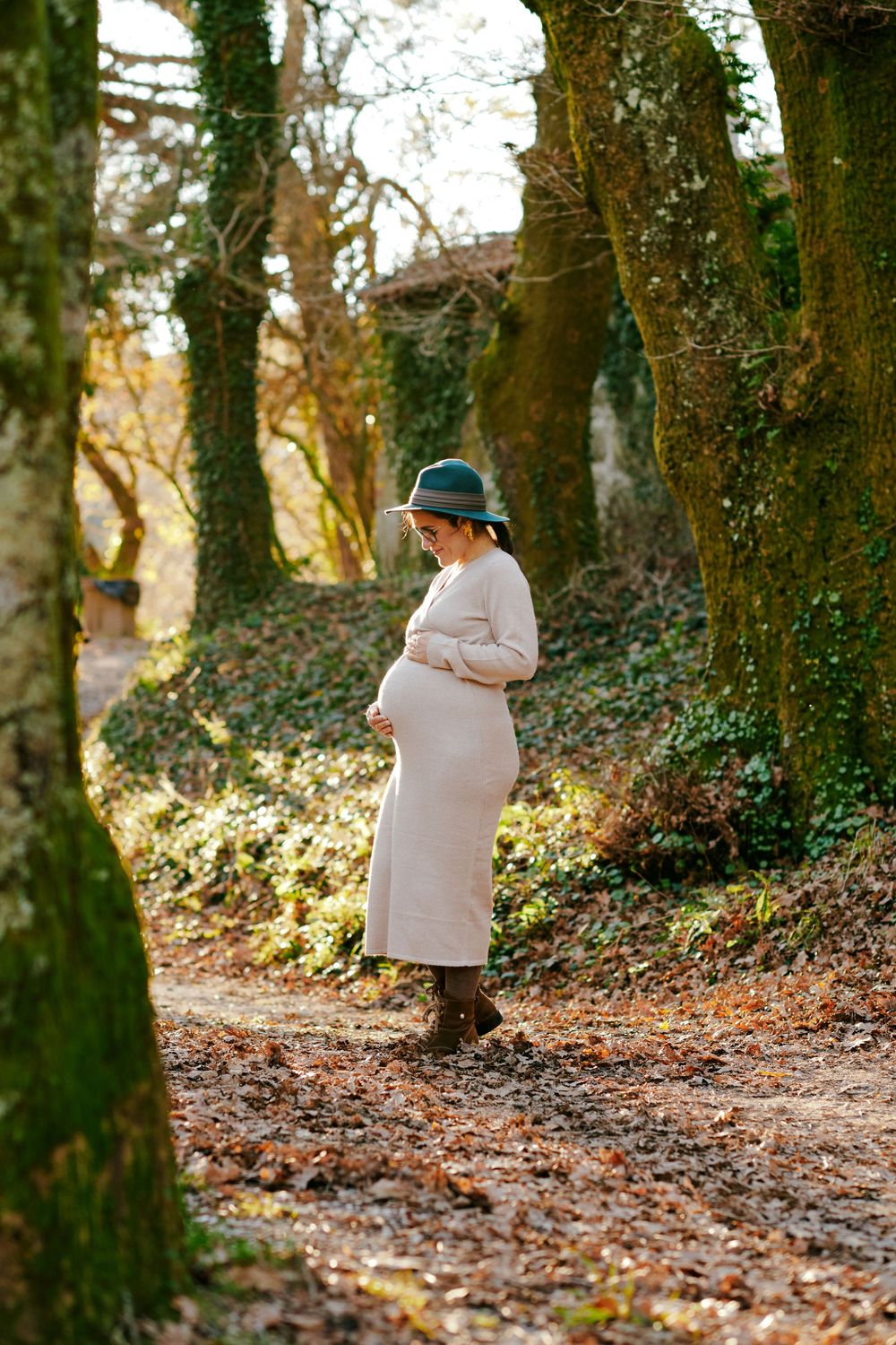 Pregnant woman in beige dress and green hat cradling belly on autumn forest path.