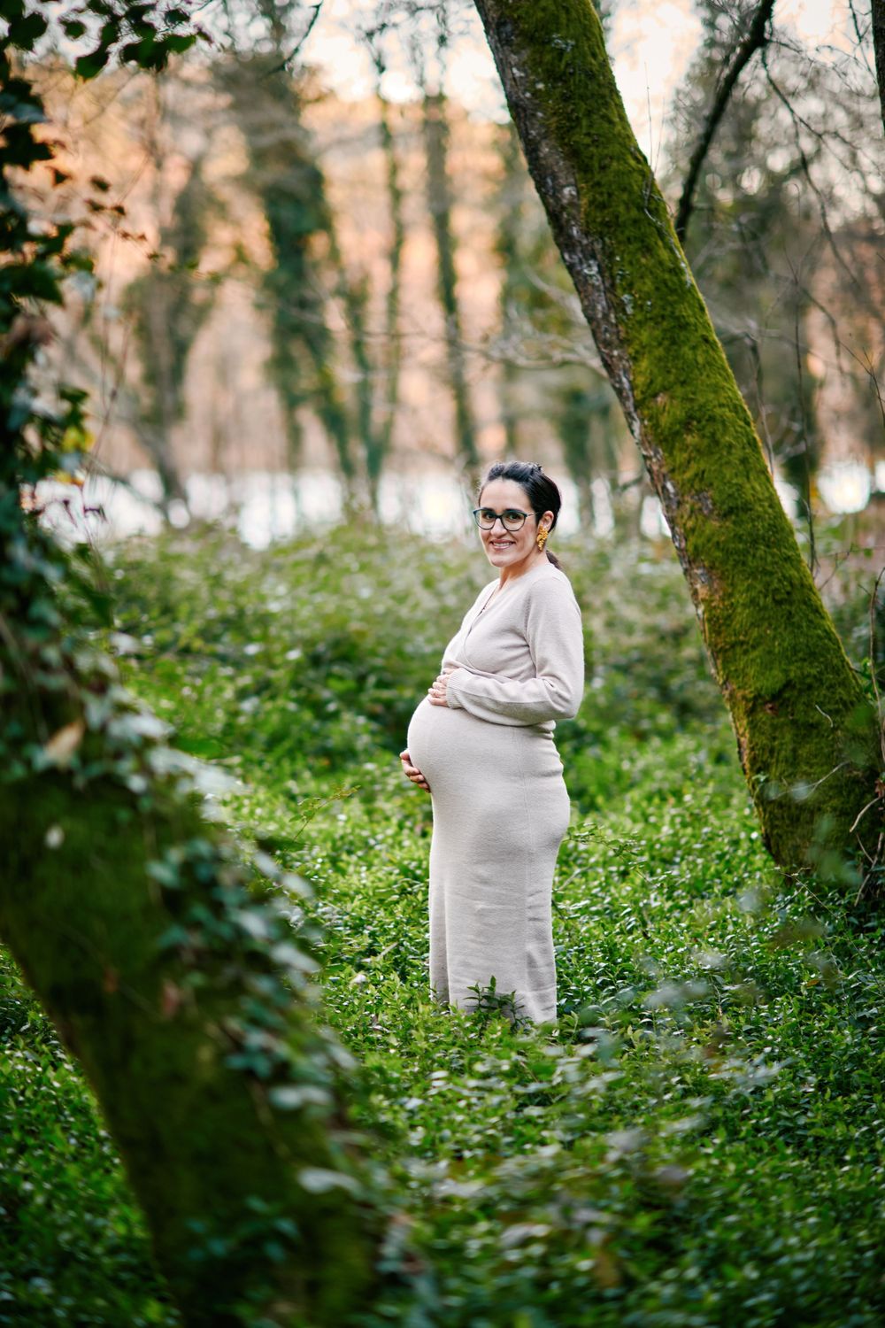 Smiling pregnant woman in beige dress holding belly among mossy trees in lush green forest.