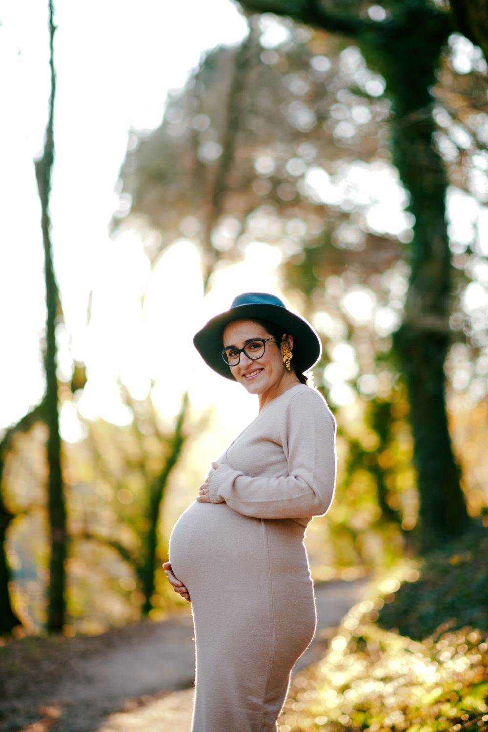 Glowing pregnant woman in beige dress and teal hat holding bump in sunlit forest.