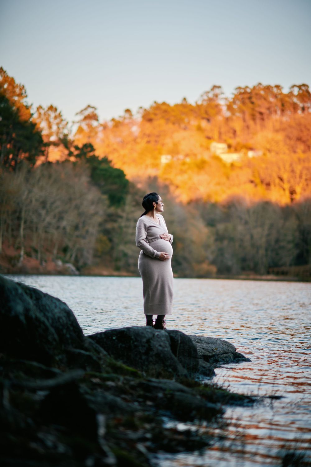 Pregnant woman in grey dress standing on lakeside rocks at golden sunset holding belly.