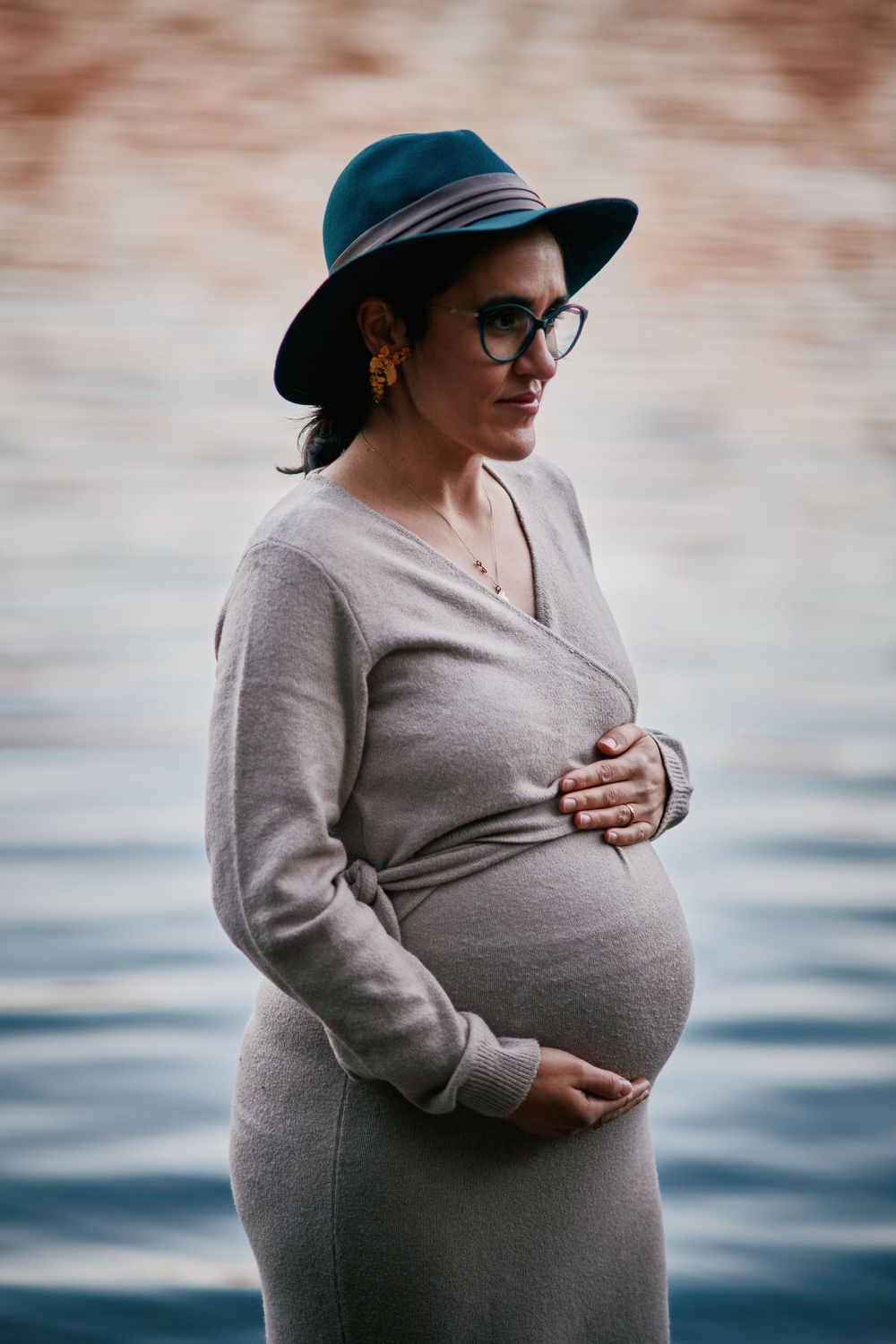 Stylish pregnant woman in grey wrap dress and teal hat cradling belly near water.