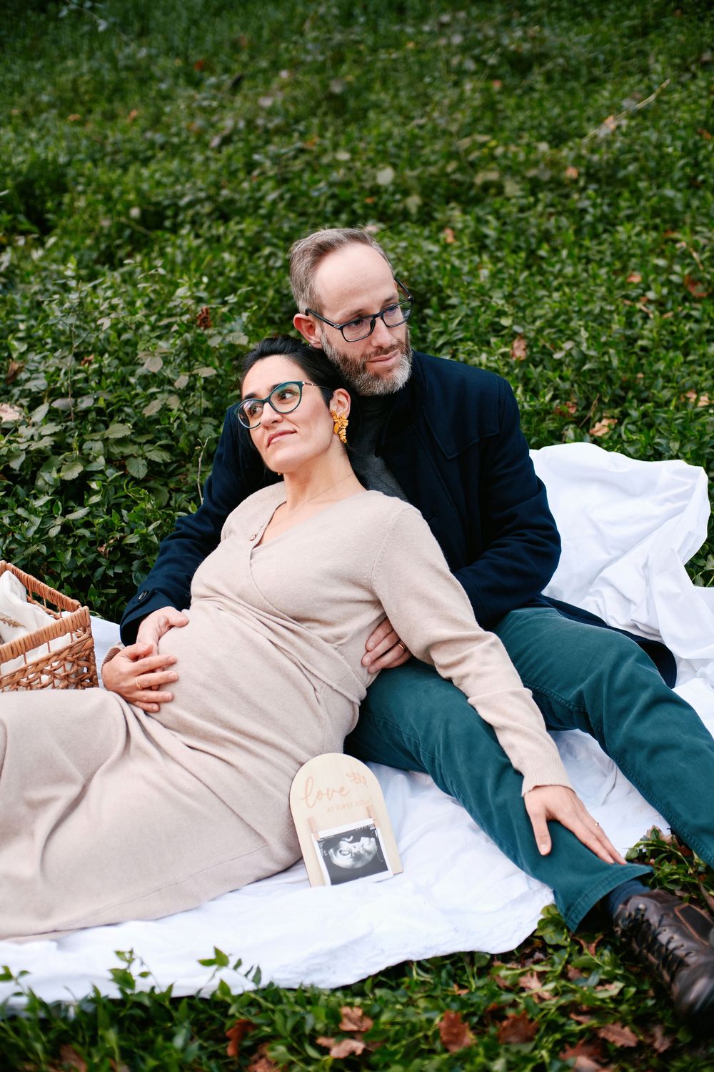 A couple relaxing on a blanket in a park, the pregnant woman leaning on her partner with an ultrasound photo.