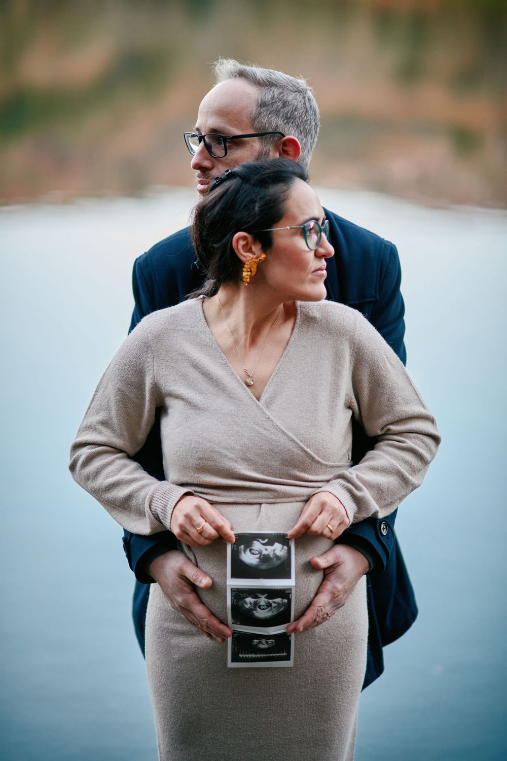 Couple holding ultrasound photos by a lake, man embracing pregnant woman from behind.