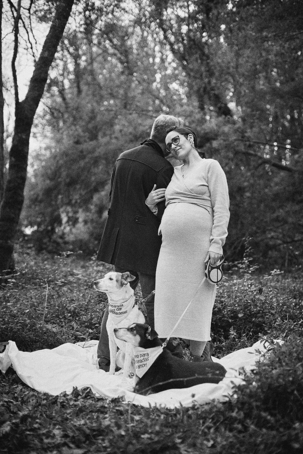 A black and white photo of a pregnant couple embracing outdoors with a dog on a blanket.