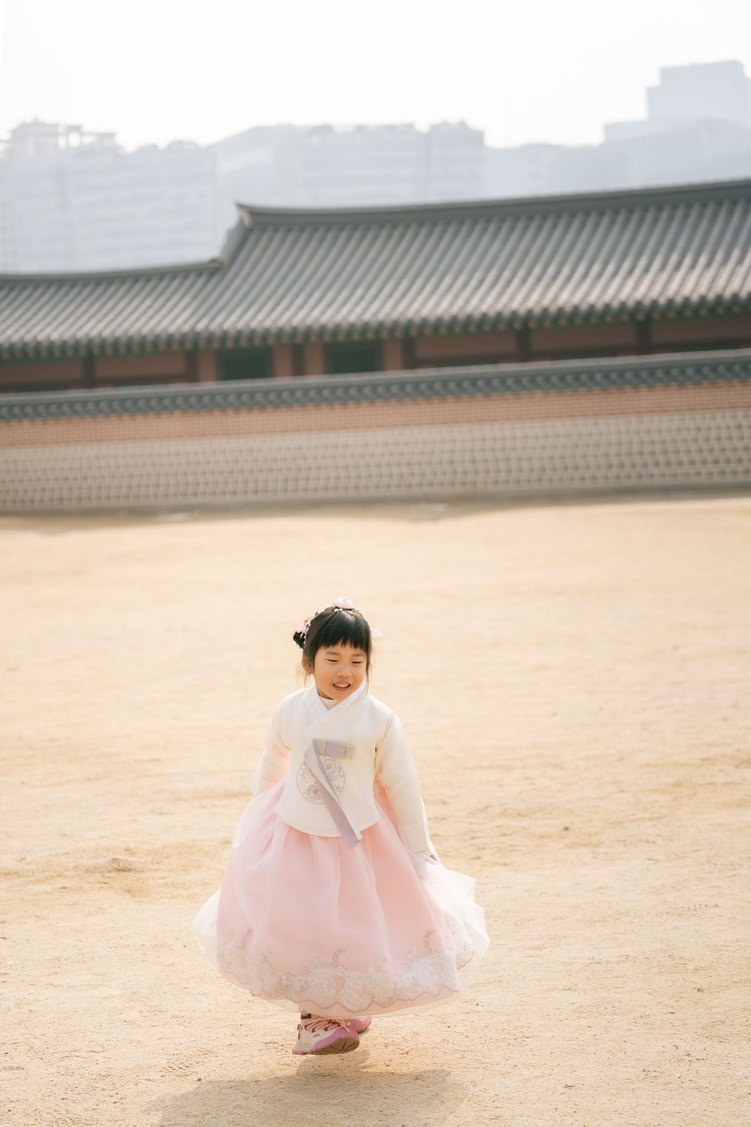 A child in a pink tulle dress crouches on sandy ground near traditional Asian architecture.