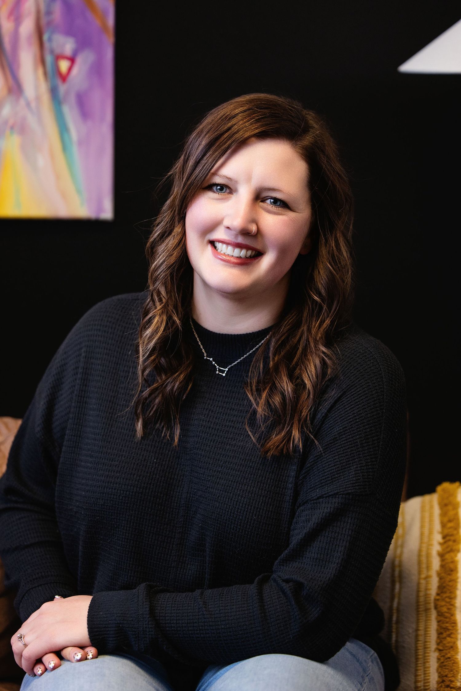 A person in a black sweater sits and smiles warmly against a dark background with colorful artwork visible.