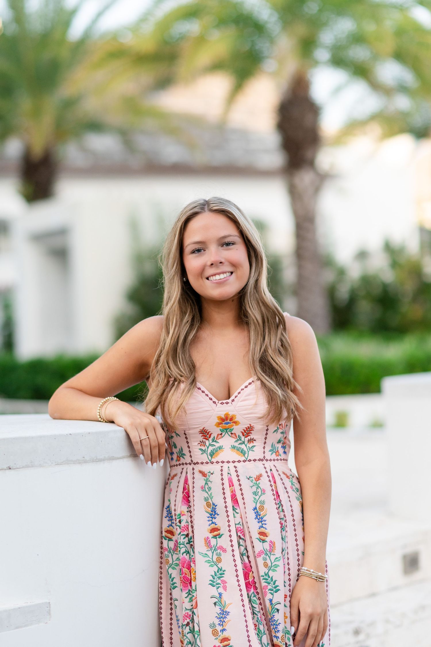 Casual outdoor photoshoot featuring a white and pink floral dress near palm trees and white walls.