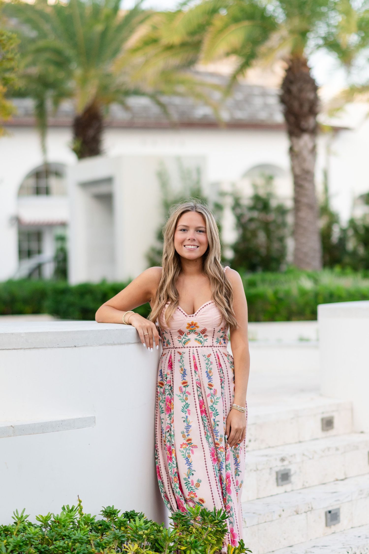 A floral sundress is worn against a backdrop of palm trees and white architecture in a tropical setting.