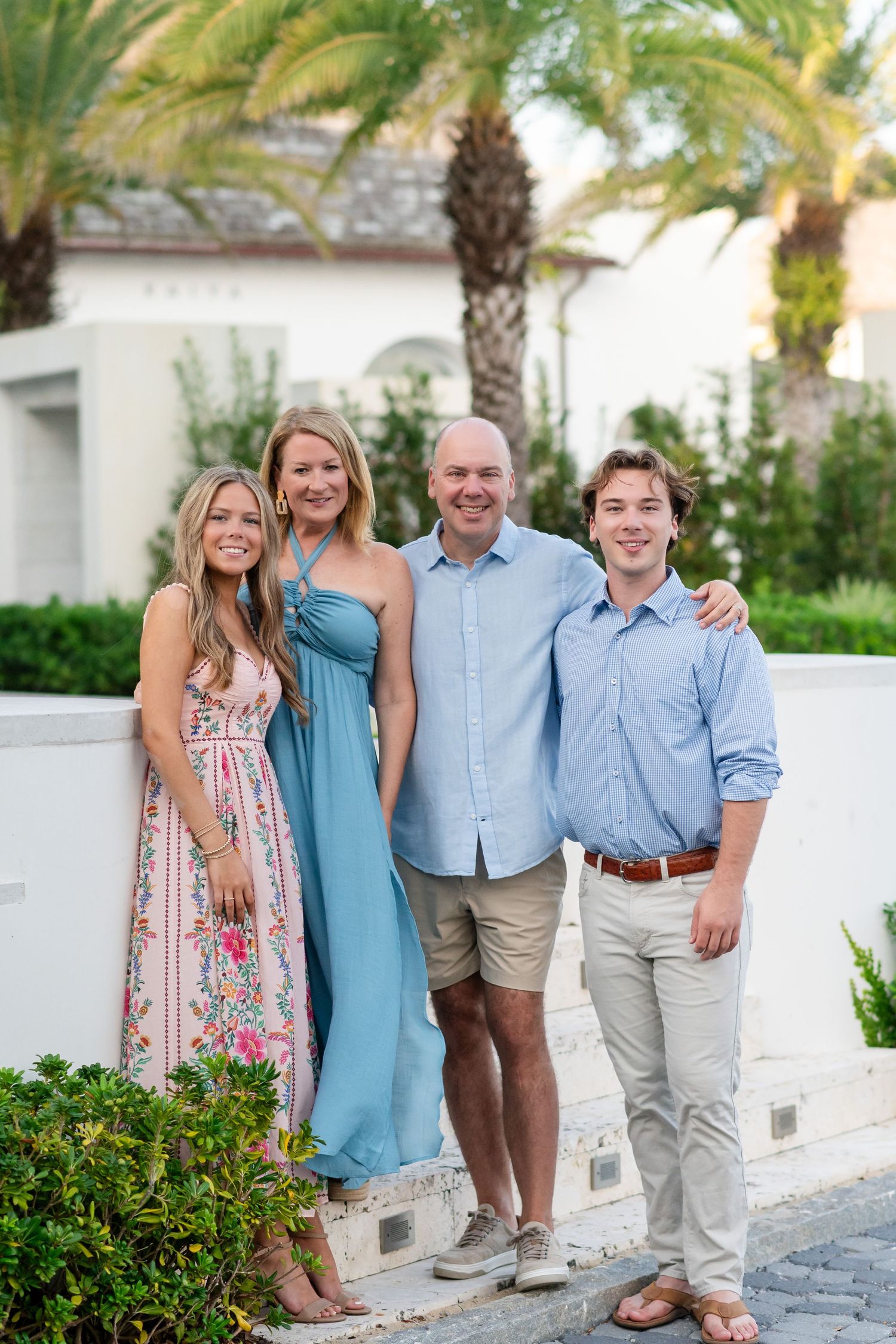 Family group poses together outdoors in coordinated blue and pink outfits against a tropical palm tree setting.