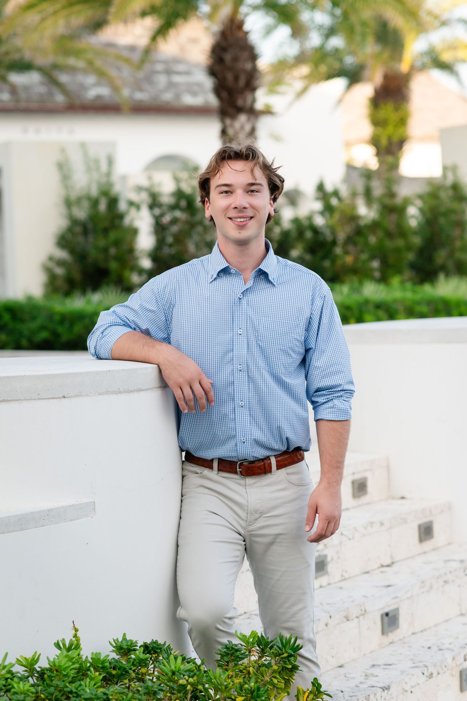 Young person in light blue button-down shirt and khaki pants poses against white wall with tropical plants.