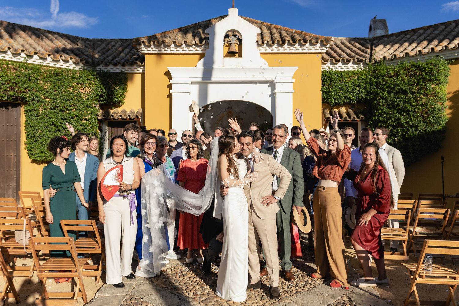 Wedding celebration group poses in front of yellow Spanish colonial style church entrance with wooden chairs.