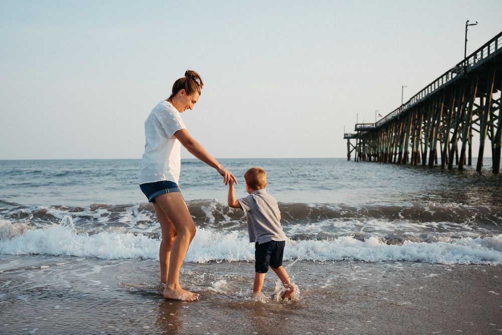A family enjoys splashing and playing together in the ocean waves near a wooden pier at sunset. Kure Beach Family Photographer