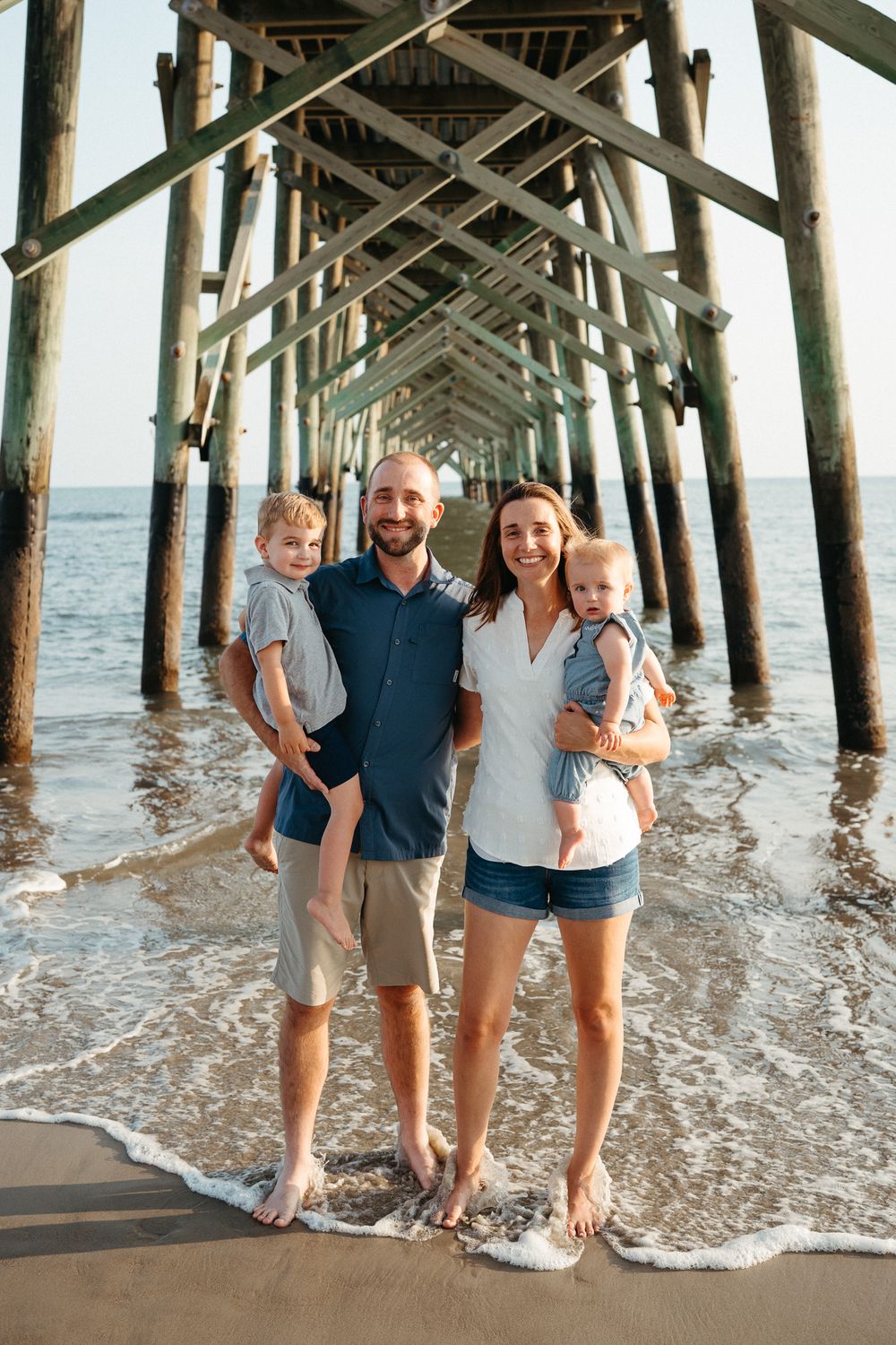 A group stands beneath a wooden pier at the beach as waves roll in during a casual photoshoot. Wilmington NC family photographer